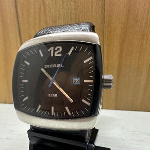 May include: A black Diesel wristwatch with a square face and silver trim. The watch face has silver hands, a date window, and the words "Diesel" and "5 bar" printed on it.