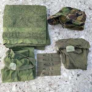 Op de afbeelding: Een set van vijf groene militaire naamschildjes met de tekst "MILLS G." gedrukt op elk schildje. De schildjes zijn in een stapel gerangschikt. Er is ook een groene handdoek, een groen T-shirt, een groen onderhemd en een camouflagepet.