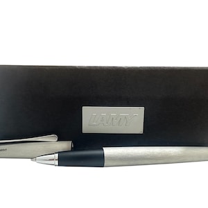 May include: A brushed silver ballpoint pen with a black grip section and a matching removable cap featuring a silver clip. The pen is displayed in front of a dark brown rectangular presentation box with a silver LAMY logo plate centered on the front.