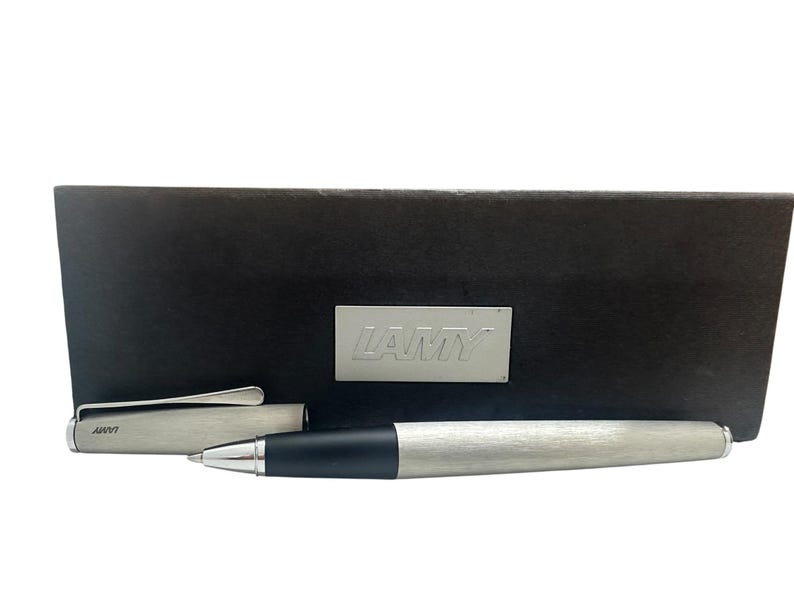 May include: A brushed silver ballpoint pen with a black grip section and a matching removable cap featuring a silver clip. The pen is displayed in front of a dark brown rectangular presentation box with a silver LAMY logo plate centered on the front.