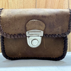 Puede incluir: Un bolso de cuero marrón con cierre de solapa y hebilla plateada. El bolso tiene un ribete cosido decorativo alrededor de los bordes.