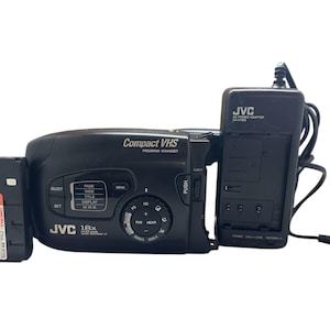 Könnte beinhalten: Ein schwarzer JVC Compact VHS Program Manager Camcorder mit einem schwarzen Netzteil. Der Camcorder hat einen LCD-Bildschirm, Tasten und ein Zoomobjektiv. Das Netzteil trägt den Text "JVC AC Power Adapter AA-V1203".