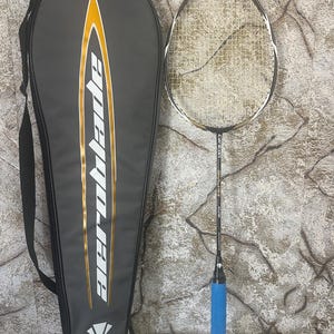 Peut inclure: Une raquette de badminton Aeroblade noire et dorée et son étui assorti. La raquette a une poignée bleue et une tête blanche et dorée. L'étui est noir avec le mot "Aeroblade" en blanc et or.