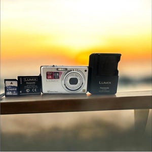 May include: A silver Lumix digital camera with a black lens, a black battery charger, a black battery, and an 8GB Kingston SD card. The camera has a 12-megapixel resolution and a 28mm wide lens. The items are arranged on a wooden surface.