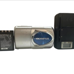 May include: A silver Olympus digital camera with a blue accent, a black battery charger, and a black battery. The camera has the brand name "Olympus" in white text. The battery has the brand name "Olympus" and other text.