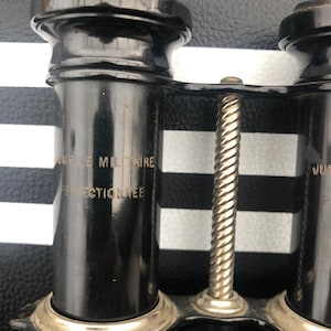 K&ouml;nnte beinhalten: Schwarzes Fernglas mit silbernem Fokussierknopf und dem Text "Jumelle Militaire Perfectionnee" an der Seite.