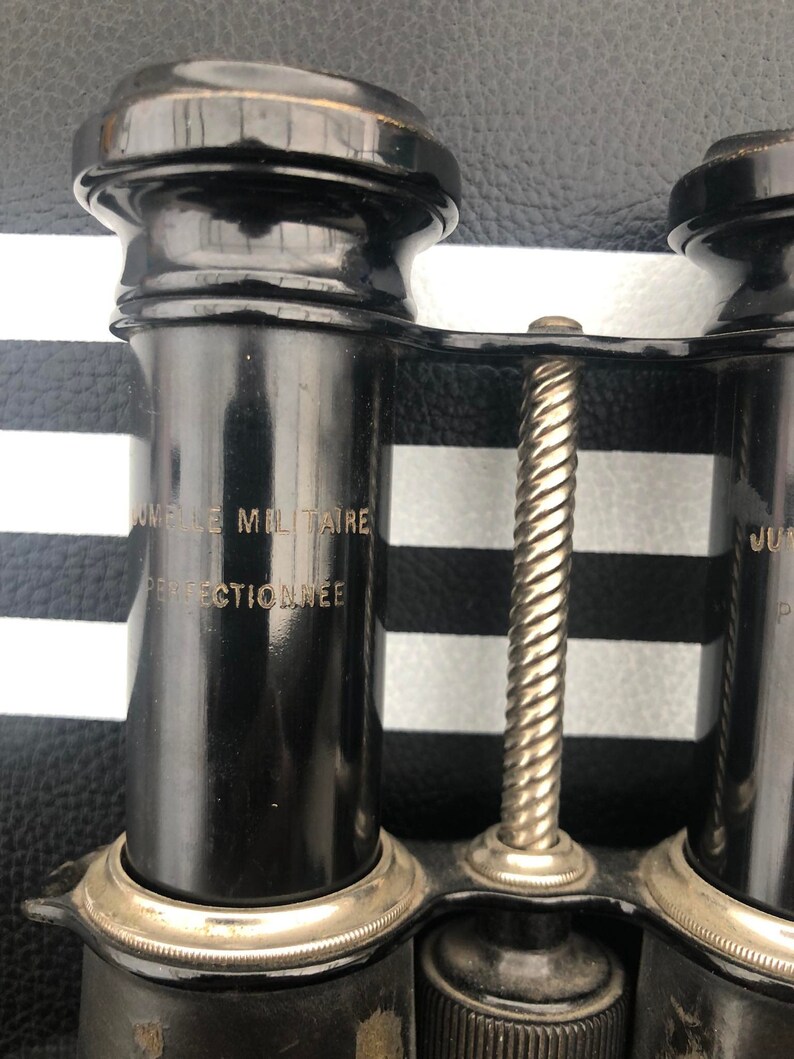 K&ouml;nnte beinhalten: Schwarzes Fernglas mit silbernem Fokussierknopf und dem Text "Jumelle Militaire Perfectionnee" an der Seite.