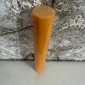 May include: A solid, cylindrical rod with a smooth, opaque, mustard-yellow finish. The object features a flat, circular top surface and tapers slightly toward the base, presenting a uniform, matte-textured appearance throughout its length.