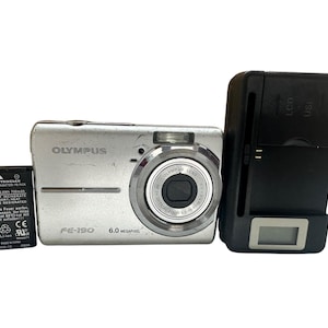 May include: Silver Olympus FE-190 6.0 megapixel digital camera with a black battery and charger. The camera has a zoom lens and the charger has an LCD and USB port.