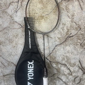 Puede incluir: Una raqueta de bádminton negra y marrón con un agarre blanco, colocada junto a una funda negra para la raqueta. La funda de la raqueta tiene el nombre de la marca "YONEX" en blanco. La raqueta tiene una red y está sobre un fondo texturizado.