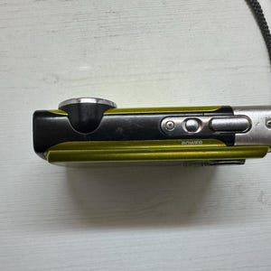 May include: A small, green and black digital camera with a silver lens and a power button. The camera has a strap attached to it.