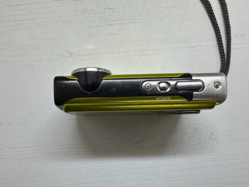 May include: A small, green and black digital camera with a silver lens and a power button. The camera has a strap attached to it.
