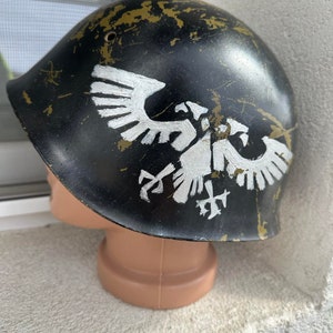 Può includere: Un elmetto militare nero con un'insegna di aquila bianca dipinta sul davanti. L'elmetto ha un aspetto usurato e rovinato con graffi e scheggiature di vernice visibili.