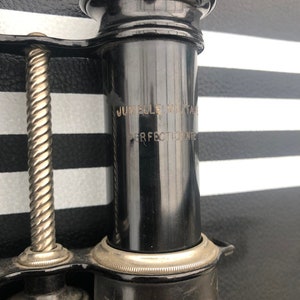 K&ouml;nnte beinhalten: Nahaufnahme eines schwarzen Vintage-Teleskops mit einem silbernen Band und der Aufschrift "Jumelle Militaire Perfectionne" an der Seite.