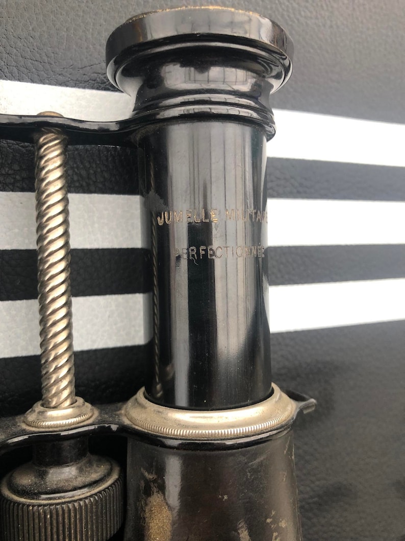 K&ouml;nnte beinhalten: Nahaufnahme eines schwarzen Vintage-Teleskops mit einem silbernen Band und der Aufschrift "Jumelle Militaire Perfectionne" an der Seite.