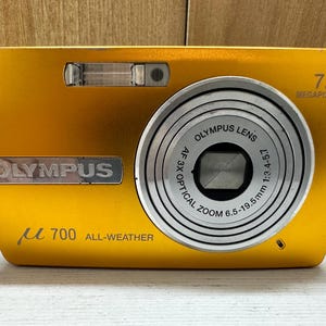 May include: A gold Olympus &micro;700 digital camera with a 7.1 megapixel resolution and a 3x optical zoom lens. The camera has a silver lens ring and a silver Olympus logo.