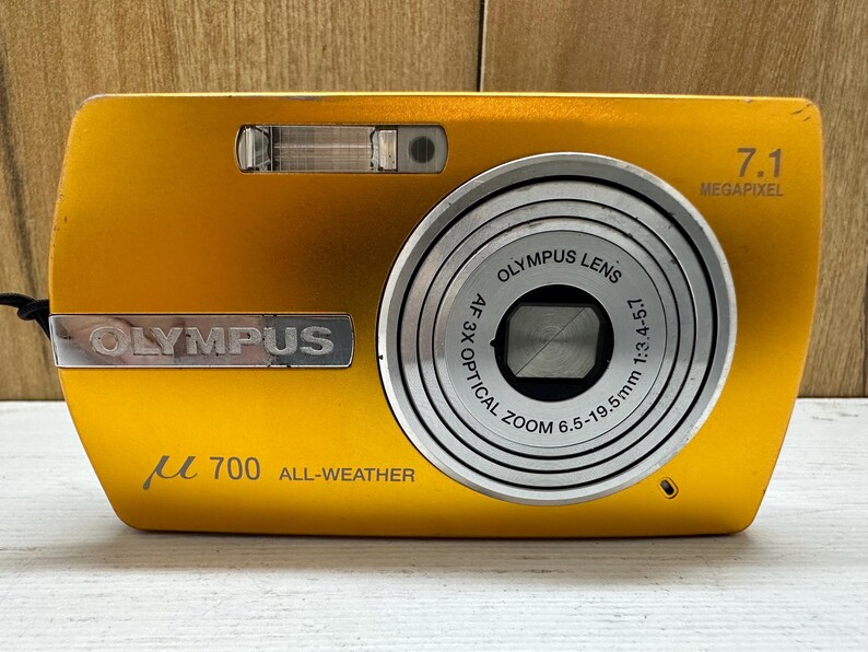 May include: A gold Olympus &micro;700 digital camera with a 7.1 megapixel resolution and a 3x optical zoom lens. The camera has a silver lens ring and a silver Olympus logo.