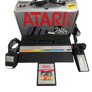 Puede incluir: Una consola de videojuegos Atari 2600 con su caja original, fuente de alimentación, joystick y un cartucho de juego. La caja es gris y roja con el logotipo de Atari y el texto "2600". La consola es negra con una franja plateada y el logotipo de Atari. El cartucho del juego está etiquetado como "Tennis".