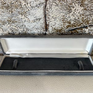 Puede incluir: Una caja de joyería rectangular abierta, con exterior de terciopelo gris e interior de satén blanco. La caja tiene un inserto de terciopelo gris oscuro con dos pequeños soportes verticales. Diseñado para guardar un collar o pulsera.