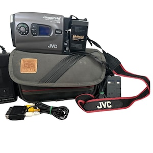 以下が含まれることがあります： 黒と赤のストラップが付いたグレーのJVCコンパクトVHSビデオカメラ、グレーのキャリングケース、黒の電源アダプター。ビデオカメラには「Compact VHS」と「JVC」の文字が印刷されています。