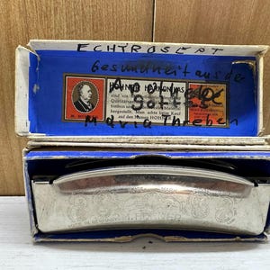 Pode incluir: Uma gaita vintage numa caixa azul e branca. A caixa tem o texto "ECHTROSEPT Gesundheit aus der HOHNER HARMONICA gott" impresso nela. A gaita é prateada com o texto "MADE IN GERMANY M. HOHNER" impresso nela.