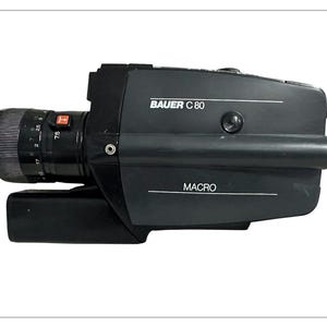 May include: A black Bauer C 80 vintage film camera with a lens and the word "MACRO" printed on the side. The lens has measurement markings. The camera is on a white background.