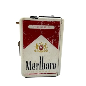 Może przedstawiać: Prostokątna metalowa papierośnica z biało-czerwonym wzorem, logo Marlboro, złotym herbem z dwoma lwami i chińskimi znakami. Powierzchnia nosi ślady użytkowania i zarysowania, z czarnym zatrzaskiem na górnej krawędzi.