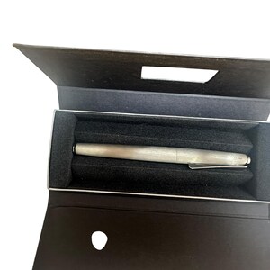 May include: A silver-toned fountain pen with a metallic finish and a pocket clip, resting horizontally inside a dark brown rectangular presentation box. The interior of the box features a black foam insert with a molded groove to hold the pen securely.