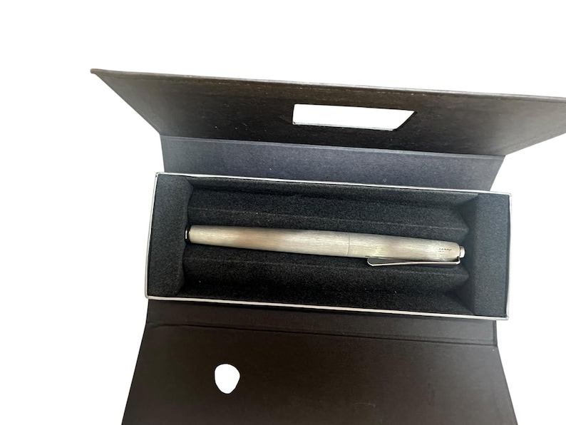 May include: A silver-toned fountain pen with a metallic finish and a pocket clip, resting horizontally inside a dark brown rectangular presentation box. The interior of the box features a black foam insert with a molded groove to hold the pen securely.