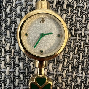 May include: A gold-tone wristwatch with a white face and green shamrock accents. The watch face has a patterned design and features a crown logo with the letters "BB".
