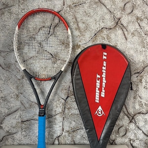 May include: A tennis racket with a red and white frame, black handle, and blue grip, next to a red and black tennis racket case. The case has the text "IMPACT Graphite Ti" printed on it. The racket is on a white surface.