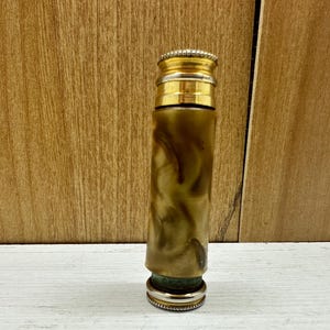 May include: A vintage perfume bottle with a gold-coloured top and base. The body of the bottle is a marbled olive green and brown. The bottle is cylindrical and stands upright on a white surface against a wooden background.