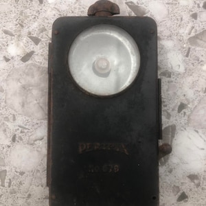 May include: A black metal flashlight with a round glass lens and a handle on the side. The flashlight has the text "PERMAX" and "No. 679" printed on it.
