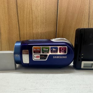 May include: A blue Samsung camcorder with a silver accent, a battery, and a black charger. The camcorder has a 42x zoom and features 16GB of memory. It also has a YouTube logo and a 3-hour battery life.