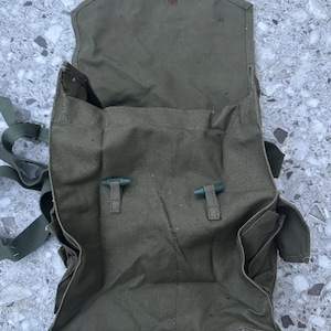 Puede incluir: Una mochila militar de lona verde con dos correas y dos hebillas. La mochila está abierta y el interior es visible. La mochila tiene un pequeño bolsillo en el lateral.