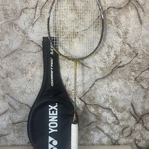 以下が含まれることがあります： 白いグリップと黒と黄色のフレームのバドミントンラケット。白い「YONEX」ロゴが入った黒いラケットケースの隣にあります。ラケットは縦に配置され、ストリングとデザインが示されています。背景はテクスチャード加工された壁です。