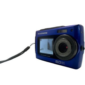 May include: A blue Maginon digital camera with a wrist strap. The camera has a screen displaying an image, a lens, and the text "8.0 MEGA PIXELS". The camera is a compact size.