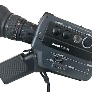 Op de afbeelding: Een zwarte Bauer S 207 XL videocamera met een zoomlens, een ingebouwde microfoon en verschillende bedieningselementen, waaronder achtergrondverlichting, fade, timer, volume, afstandsbediening en monitor.