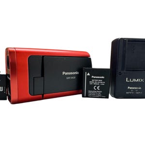Może przedstawiać: Czerwona kamera Panasonic SDR-SW20 z czarnym obiektywem i gniazdem na kartę pamięci. Kamera jest wyposażona w czarny akumulator Panasonic, czarną ładowarkę do akumulatora Panasonic i dwie karty pamięci.
