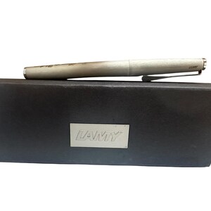 May include: A silver-toned Lamy fountain pen with a brushed metal finish and a pocket clip, resting horizontally on a rectangular black presentation box that features a silver-colored metal plaque with the brand name LAMY embossed on the front.
