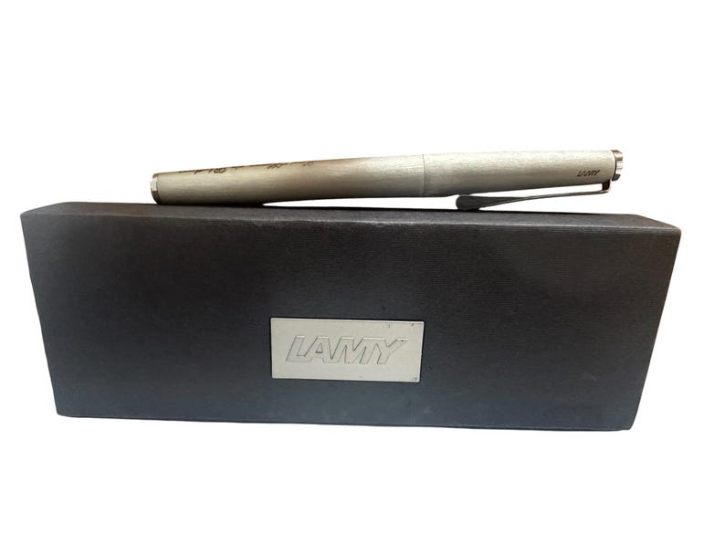 May include: A silver-toned Lamy fountain pen with a brushed metal finish and a pocket clip, resting horizontally on a rectangular black presentation box that features a silver-colored metal plaque with the brand name LAMY embossed on the front.