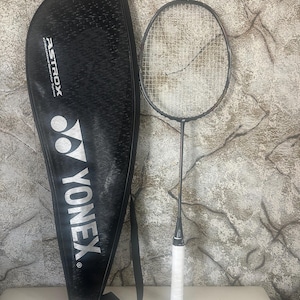 Può includere: Una racchetta da badminton nera con un'impugnatura bianca accanto a una custodia nera. La racchetta ha un telaio nero e una rete a maglie. La custodia porta la scritta "YONEX" in bianco e la scritta "ASTROX".