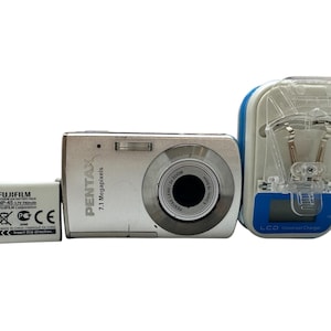 Könnte beinhalten: Eine silberne Pentax Digitalkamera mit 7,1 Megapixeln, einem Fujifilm Akku und einem Universal-Ladegerät. Die Kamera hat ein Zoomobjektiv. Das Ladegerät ist weiß und blau.