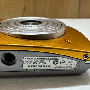 May include: A gold digital camera with a silver lens and a black label that reads "OLYMPUS IMAGING CORP. DIGITAL CAMERA MODEL NO. &mu; 700 E B75526918 D33235 DC 3.7V MADE IN CHINA".