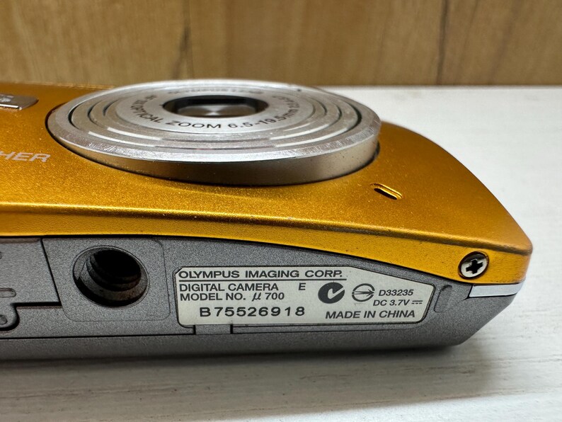 May include: A gold digital camera with a silver lens and a black label that reads "OLYMPUS IMAGING CORP. DIGITAL CAMERA MODEL NO. &mu; 700 E B75526918 D33235 DC 3.7V MADE IN CHINA".