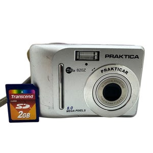 May include: Silver Praktica DPix 820Z digital camera with a Prakticar lens and a 2GB Transcend SD card. The camera has a built-in flash and the text "8.0 MEGA PIXELS".