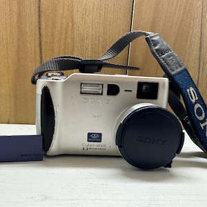 May include: A silver Sony Cybershot digital camera with a black lens cap and a blue and navy blue strap. A blue memory stick is inserted into the camera. The camera has a 3.3 megapixel resolution.