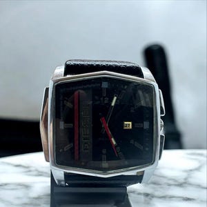 May include: A Diesel wristwatch with a black face, silver case, and black leather band. The watch features a red accent stripe and the brand name "DIESEL" vertically down the face. The date is displayed at the 3 o'clock position.
