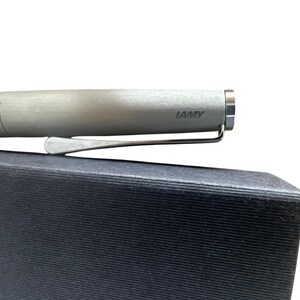 May include: A brushed silver Lamy fountain pen with a metallic finish and a silver-toned pocket clip. The brand name Lamy is printed in dark lettering on the side of the cap. The pen is positioned horizontally against a dark, textured surface.