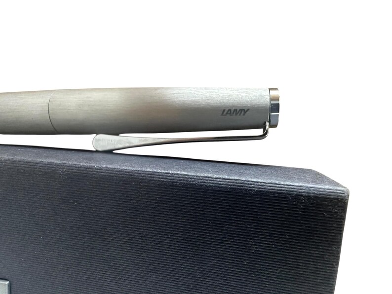 May include: A brushed silver Lamy fountain pen with a metallic finish and a silver-toned pocket clip. The brand name Lamy is printed in dark lettering on the side of the cap. The pen is positioned horizontally against a dark, textured surface.